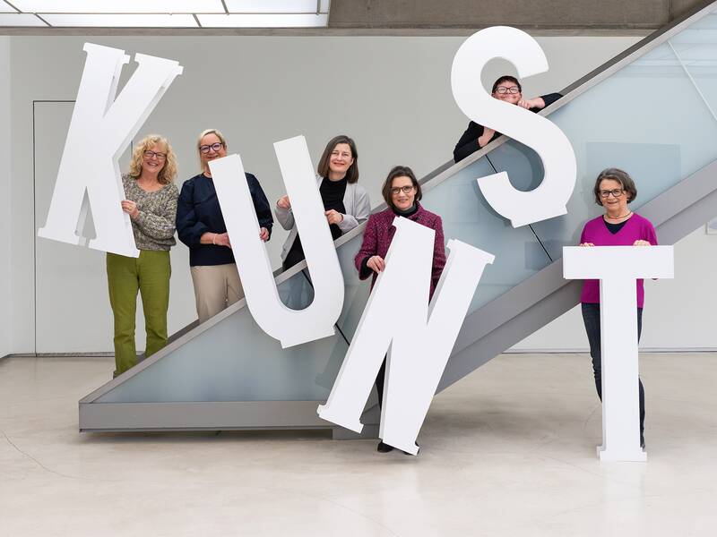 Sechs Menschen stehen in der Städtischen Galerie und halten große weiße Buchstaben, die zusammen das Wort "KUNST" bilden.