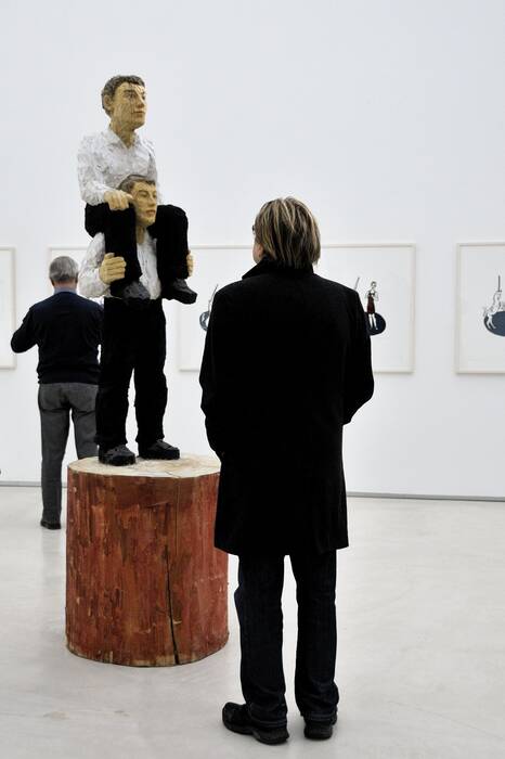 "huckepack", 1998 von Stephan Balkenhol. Bronzeskulptur zeigt zwei Figuren mit weißem Hemd und schwarzer Hose. Die eine Figur trägt die andere Figur auf dem Rücken. Ein Ausstellungsbesucher steht davor.