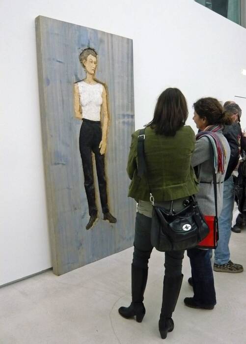 Ausstellungsbesucher vor einem großformatigen Holzrelief "Frau", 1999 von Stephan Balkenhol. Die dargestellte Frau trägt ein weißes Oberteil, eine schwarze Hose und schwarze Schuhe. Der Hintergrund ist in einem blassen Blauton angestrichen.