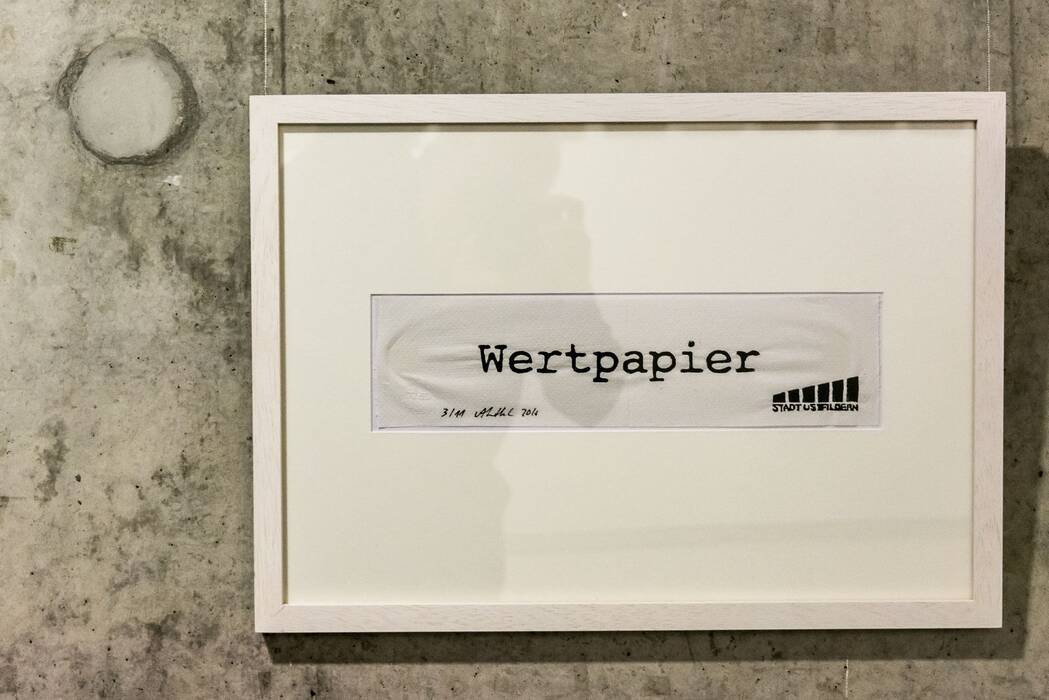 "Wertpapier Ostfildern", 2016 von Anja Luithle. In einem weißem Rahmen mit Passepartout befindet sich ein Schriftzug und das Ostfildener Logo.
