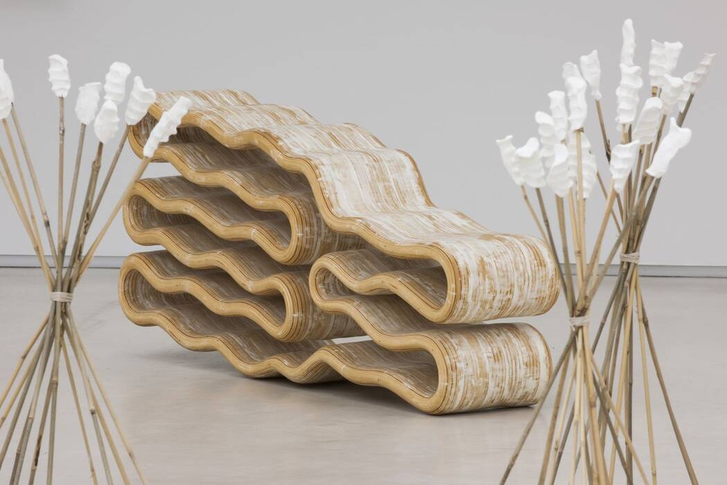  Wellenförmige Holzskulptur von Mélanie Lachièze-Rey. Sie besteht aus mehreren übereinanderliegenden, geschwungenen Schichten aus hellem Holz mit weißen Farbakzenten. Im Vordergrund befinden sich zwei Bündel dünner, bambusartiger Stäbe.