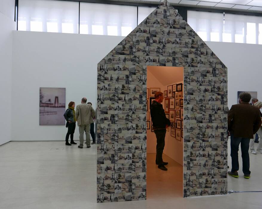Installation von Gert Wiedmaier in Form eines Hauses. Die Außenwand ist mit lauter schwarzweiß Bildern bedeckt. Durch die offene Türaussparung sieht man einen Besucher vor kleinformatigen Fotoarbeiten stehen. Im Hintergrund weitere Besucher vor einer Wachsarbeit mit urbanem Motiv.