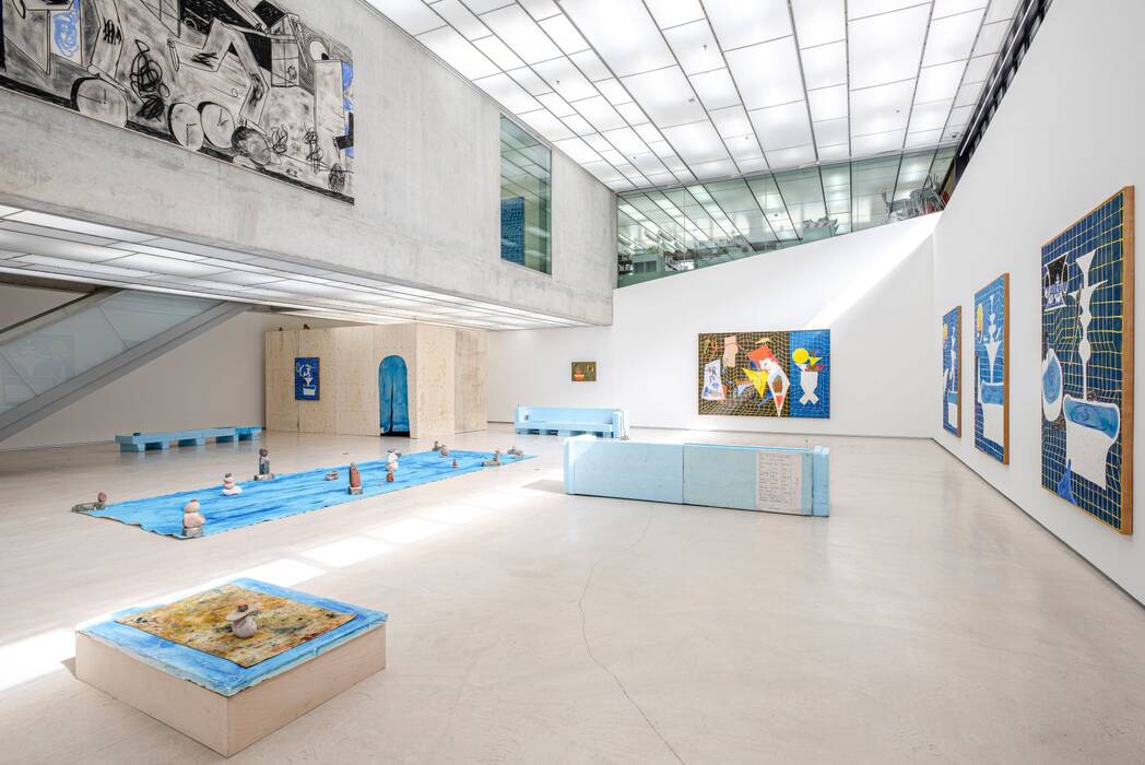 Ausstellungsansicht "Keine Zeit zum Baden" von Jan Zöller. Große Gemälde in Blau- und Gelbtönen dominieren die Wände. Auf dem Boden ist eine Installation zu sehen: Eine blaue Plane, auf dem kleine Steinskulpturen angeordnet sind. Im Vordergrund steht ein quadratisches Objekt mit blau, gelber Oberfläche, auf der wiederum vier Steine aufeinander gestapelt sind.
