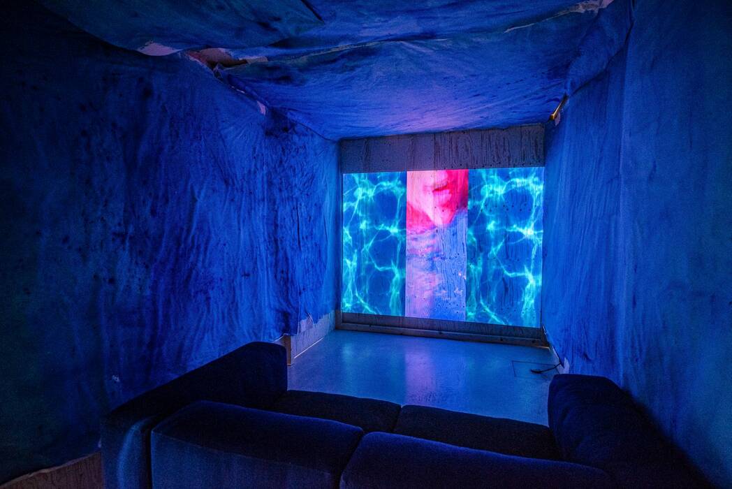 Ausstellung Jan Zöller: Ein kleiner Raum mit blauem Stoff ausgekleidet. An einer Wand ist eine Projektion zu sehen, die Wasser zeigt. Im Raum steht eine dunkelblaue Couch. Die Szene ist in dunklen Blautönen und Pink gehalten.