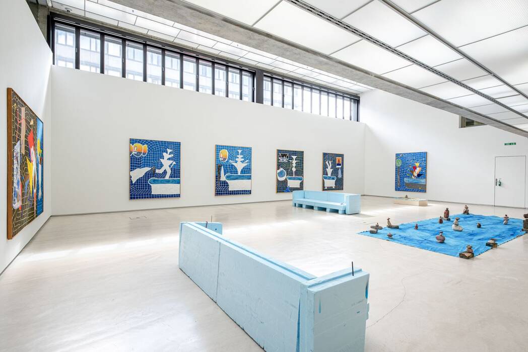 Ausstellungsansicht Jan Zöller "keine Zeit zum Baden". An den Wänden dominieren großformatige Malereien aus der Werkgruppe "Badebrunnen". Zwei Sofaähnliche Gebilde in hellblau stehen im Raum. Auf dem Boden eine blaue Plane mit gestapelten Steinen.