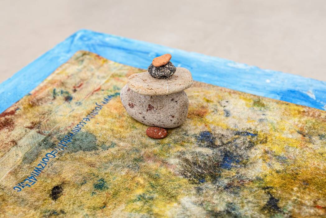 Ausstellung von Jan Zöller: Detailansicht. Fünf aufgestapelte Steine in den Farben Braun, Ocker, Rot, Orange. Untergrund in Erdtönen. Eine blaue Ecke umrahmt die Szene.