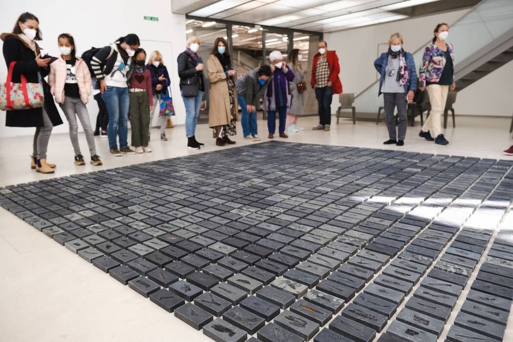 Schwarze Tafeln sind zu einem Rechteck angeordnet. Installation "Ashes to Ashes" von Jeewi Lee. Ausstellungsbesucher stehen drumherum.