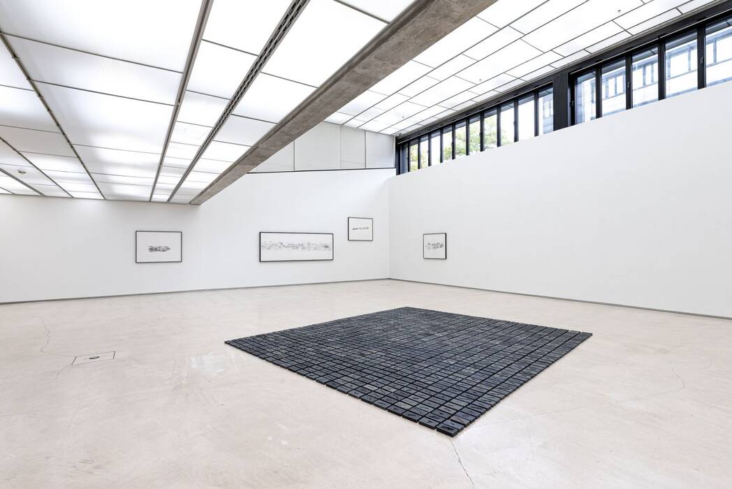 Ausstellung "Revision" von Jeewi Lee. Im Zentrum des Raumes befindet sich eine Installation mit schwarzen Tafeln. An der Wand hängen vier minimalistische Bilder in schwarzen Rahmen.