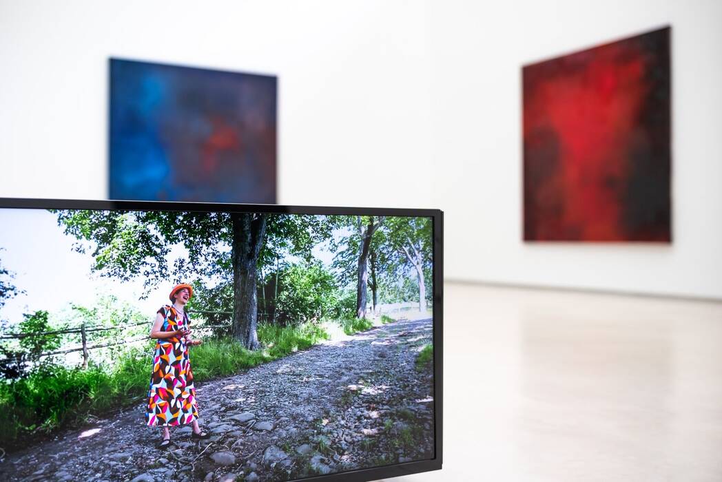 Ein Bildschirm zeigt eine Frau mit Hut in einem bunten Kleid auf einem Feldweg. Hintergrund: Eine blaue und eine rote quadratische Malerei. Ausstellung "Thinking About Video Art, Kunst am Wegesrand" von Anette C. Halm