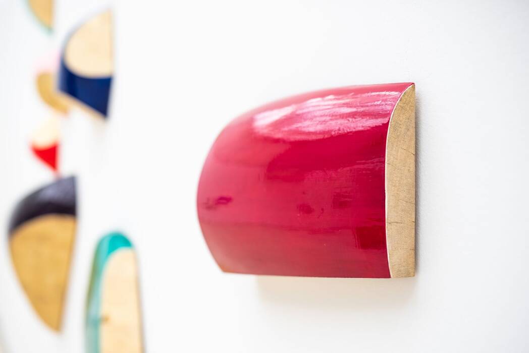 Günther Holder, Wandarbeiten.  Das Bild zeigt eine Nahaufnahme einer Reihe von halbrunden Wandobjekten auf einer weißen Wand. Das Objekt im Vordergrund ist ein leuchtend rotes, glänzendes Halbrund.