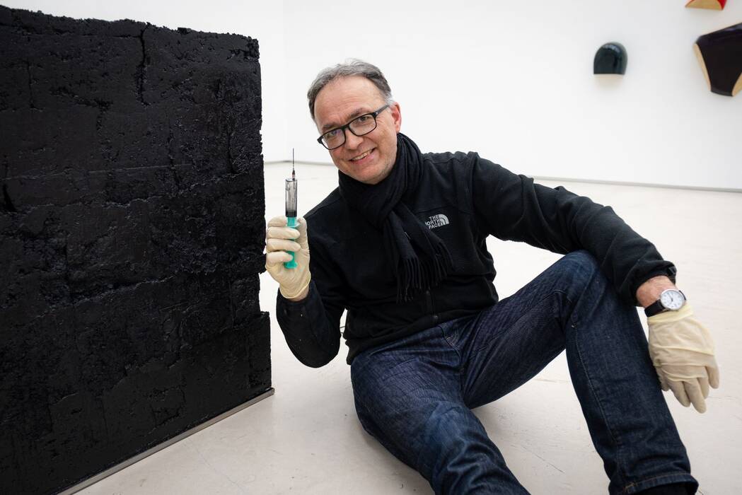 Matthias Lutzeyer bei der Arbeit. Er hält eine Spritze in die Höhe und sitzt neben einem schwarzen Kubus. 