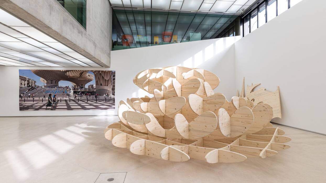 Ausstellung "J. MAYER H." An der Wand hängt ein Großformat einer modernen, städtischen, organisch geformten Architektur. Rechts steht eine große, organisch geformte Holzskulptur. 
