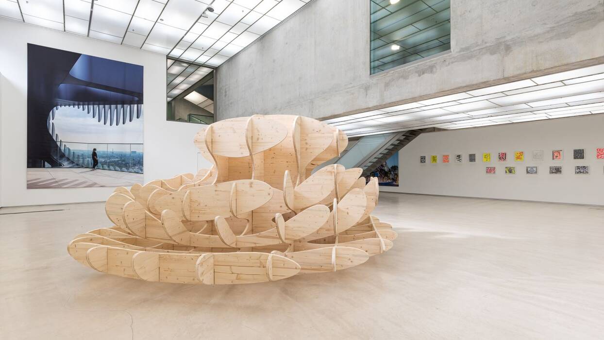 Ausstellungsansicht "Maßstäbe" von J.Mayer.H. Eine große, organisch geformte Holzskulptur steht prominent im Raum. Ein großes, wandflächiges Plakat zeigt eine moderne, städtische Architektur. Im Hintergrund erkennt man eine Reihe von Collagen in grellen Farben.