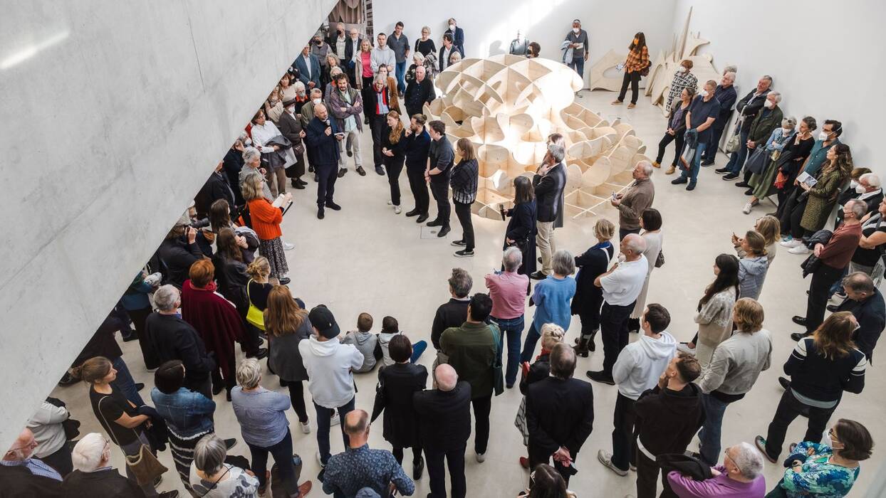 Blick von oben in den Galerieraum. Der Stararchitekt beantwortet die Fragen der Student*innen. Hinter ihnen eine Holzskulptur. Drumherum die Vernissagebesucher.