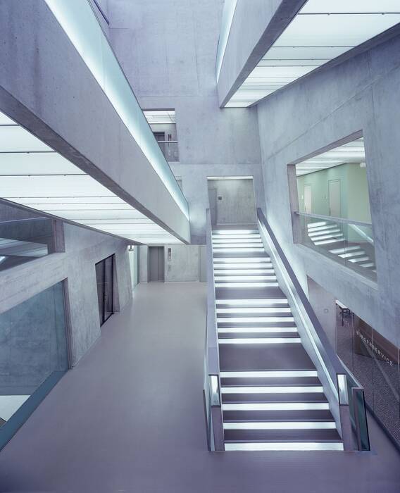 "Stadthaus", Innenansicht, vom Architketen J.MAYER.H. Im Mittelpunkt steht eine Treppe aus Beton mit integrierter Beleuchtung. Es sind mehrere Ebenen und Gänge aus Beton und Glas zu sehen, die sich ineinander verzahnen. Die gesamte Komposition ist geprägt von klaren Linien und geometrischen Formen.
