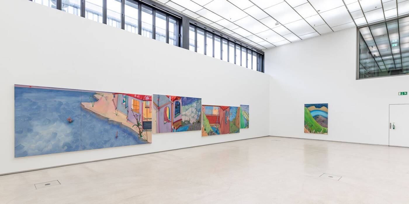 An der großen Galeriewand hängen wellenartig und leichtversetzt großformatige Exponate. Sie zeigen Gebäudefragmente in Verbindung mit Meer, Felsen und Hügeln. Ausstellung "Zu viel Blau wird schnell Nacht" von Fabian Treiber.