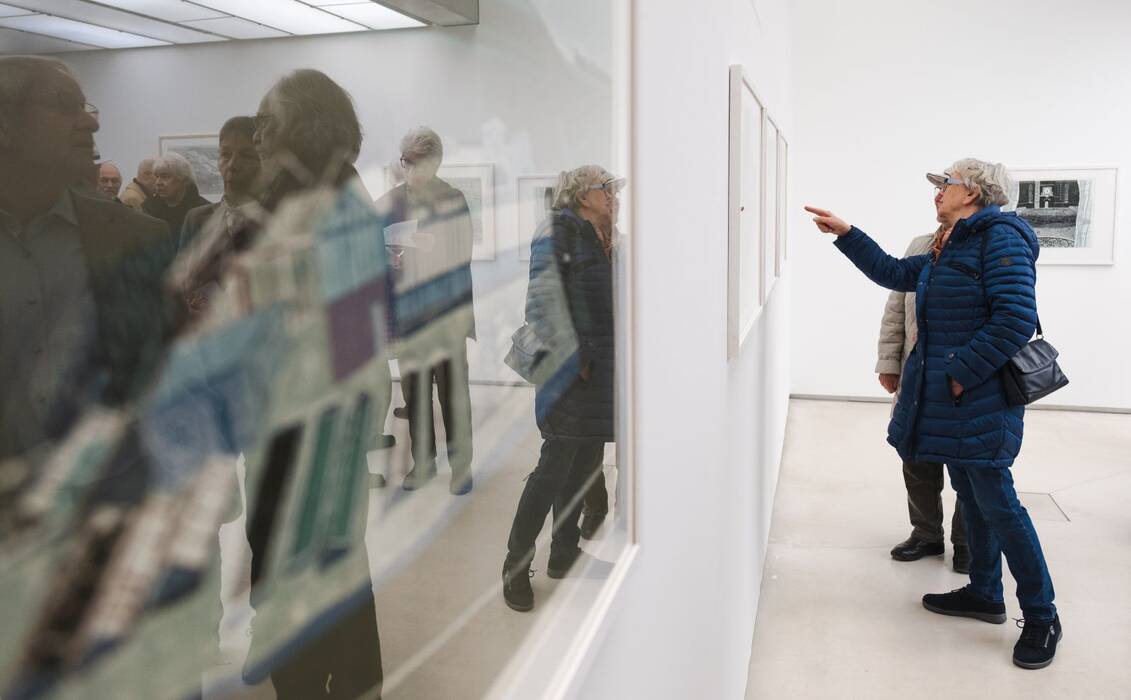 Ausstellungsbesucher betrachten einen gerahmten Linolschnitt. Im Vordergrund das Spiegelbild der Besucher.
