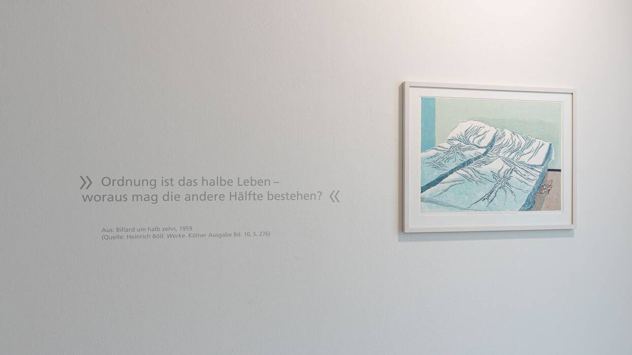 Bett mit zerknitterten Laken in den Tönen Weiß, Hellblau, Blau. Rote Damenschuhe stehen davor. Links ein Zitat: Ordnung ist das halbe Leben, - woraus mag die andere Hälfte bestehen?