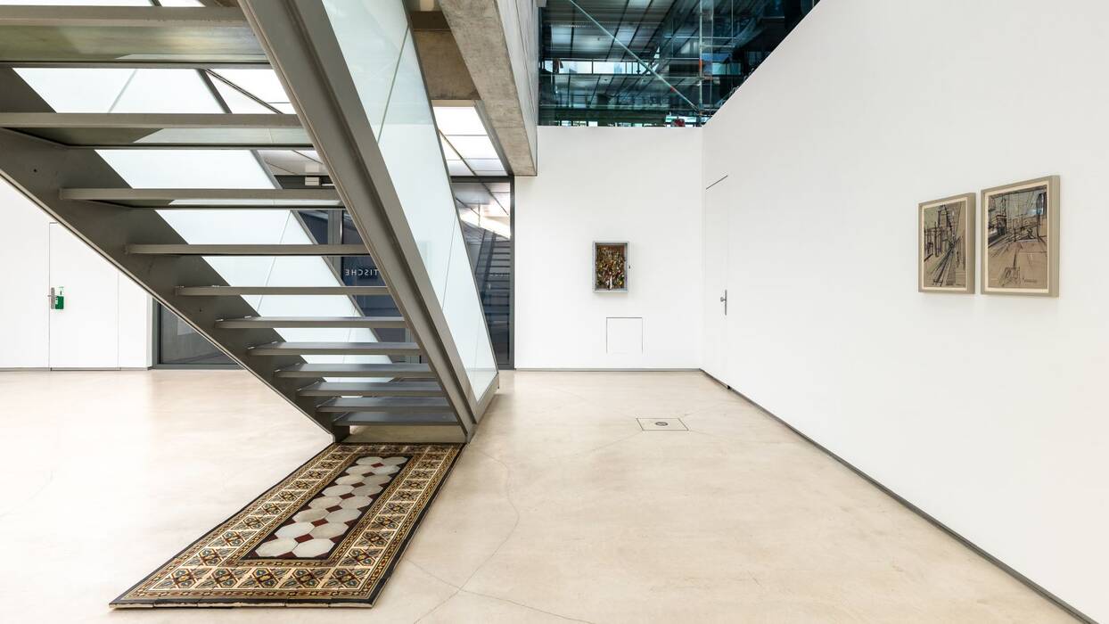 Keramikfliesen unter der Treppe mit bunten, floralen Mustern um eine Hexagon-Reihung herum. An der Wand hängen zwei Zeichnungen in Schwarz auf braunem Papier.