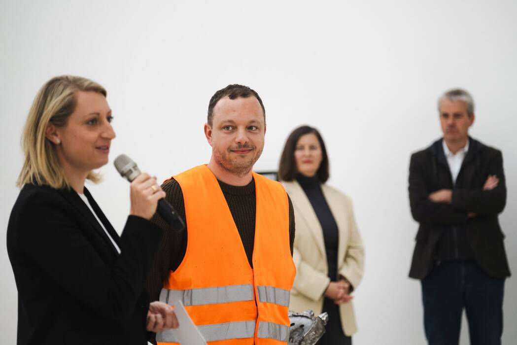 Im Fokus steht ein Mann, der Künstler Erik Sturm, in Warnweste. Die Kuratorin der städtischen Museen Heilbronn/Kunsthalle Vogelmann Carolin Wurzbacher spricht neben ihm ins Mikrofon. 