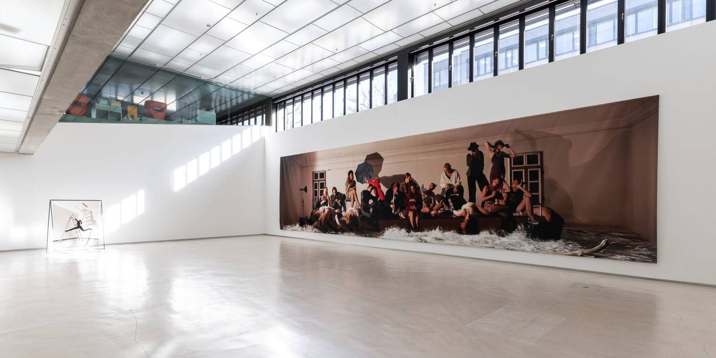 Hauptwerk der Ausstellung: Untergang der Méduse, farbiger Inkjet-Print auf PVC-Plane, 300 x 1200 cm, 2023/2024. Die dargestellten Personen sitzen, liegen oder stehen und scheinen in verschiedenen Aktivitäten beschäftigt zu sein. Im Hintergrund ein aufgestellter Rahmen mit einer schwarzweiß Fotoarbeit "sicher/unsicher".