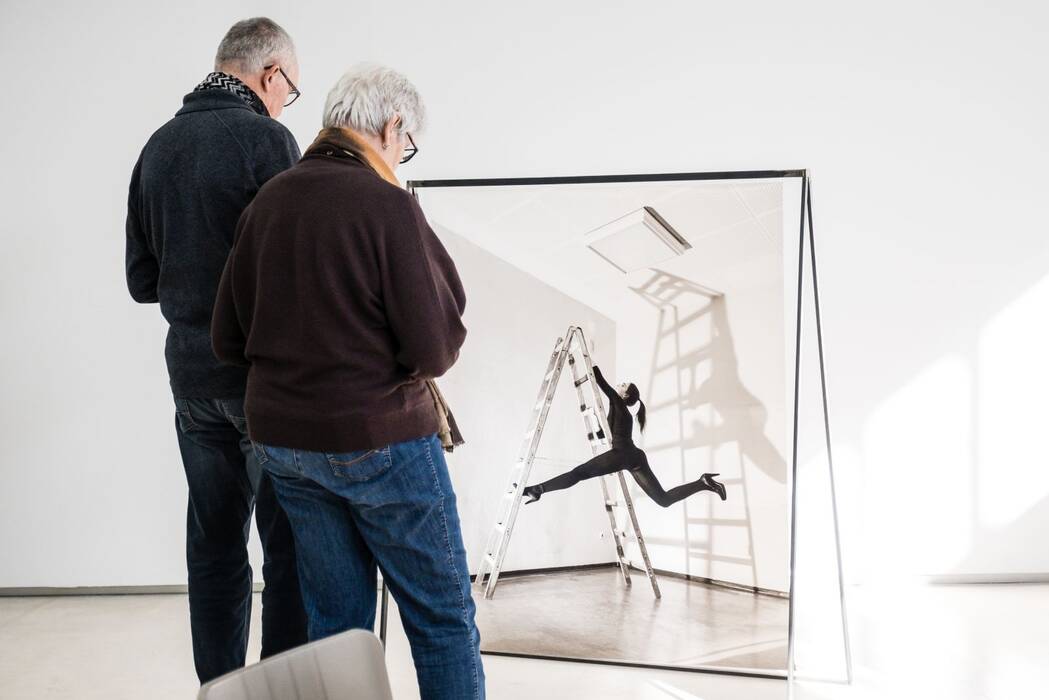 Besucher vor der Serie "sicher/unsicher", schwarzweiß Fotografie von Hannah J. Kohler. Eine schwarzgekleidete Frau mit Pferdeschwanz und Pumps hält sich an einer Stehleiter fest und streckt die Beine. 