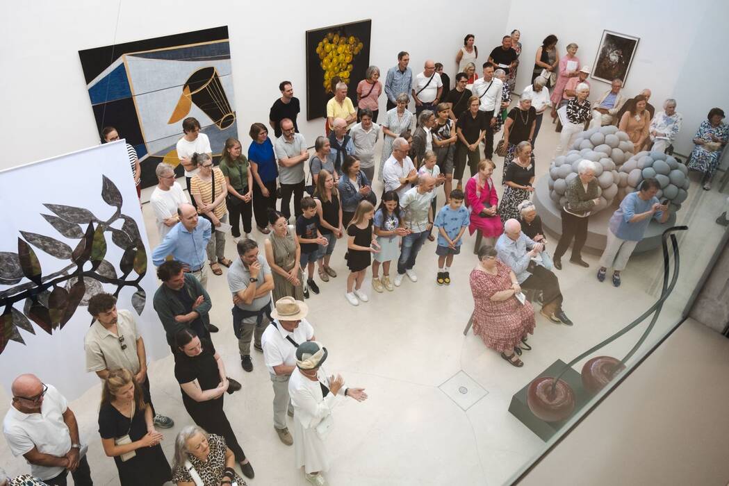 Blick von obenn ins Untergeschoss: Der mit vielen Menschen angefüllter Galerieraum. Im Vordergrund riesige rote Kirschen. Im Hintergrund farbige Bilder an den Wänden. 