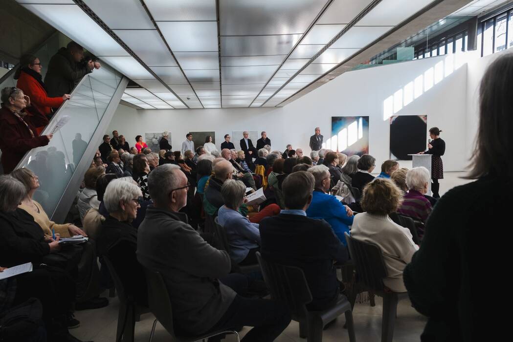 Publikum lauscht der Eröffnungsrede von Hannah Eckstein bei der Eröffnungsveranstaltung, im Hintergrund Werke von Philipp Haager