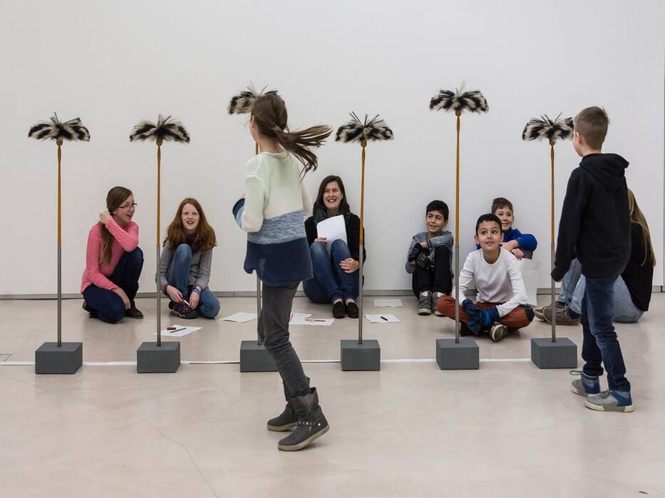Kunstvermittlung der Kunst-Scouts in der Ausstellung "schöne Aussichten" von Anja Luithle, 2016. Sechs graue, quadratische Sockel halten Stiele, auf deren Ende Schwarzweiße "Putzwedel" stecken. Die Galerieleiterin sitzt mit lachenden Kindern dahinter. Zwei Kinder stehen mit dem Rücken zum Betrachter.