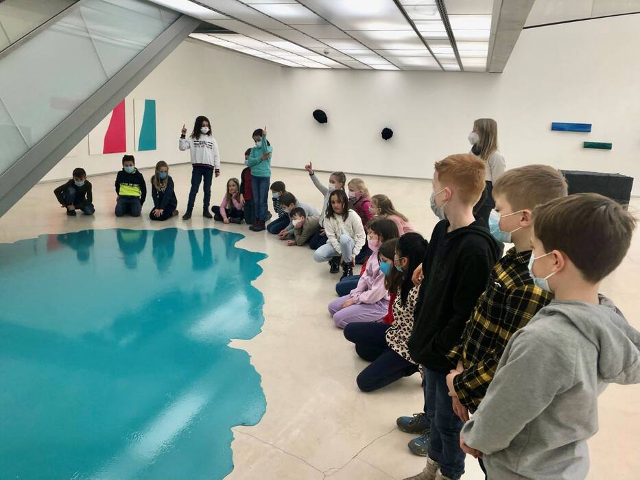 Schulklasse während eines Workshops "Auge blick mal". Die Kinder stehen und sitzen um einen türkisfarbenen Farbfleck auf dem Boden.