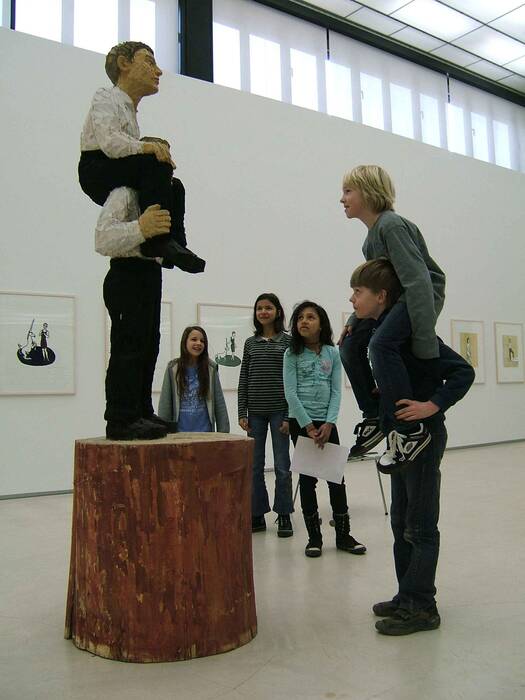 Kinder stehen vor der Holzskulptur "huckepack", 1998 von Stephan Balkenhol. Die Skulptur stellt zwei Figuren in weißem Hemd und schwarzer Hose auf einem Holzsockel dar, die einander huckepack tragen. Zwei Kinder ahmen die Figuren nach. 