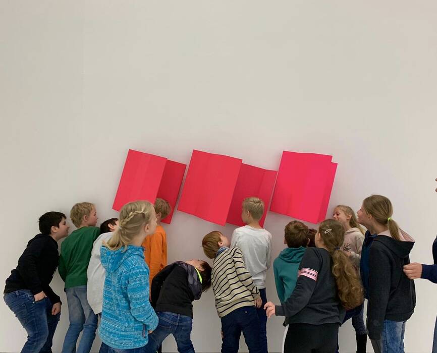 "Veränderung", Lack auf Aluminium, 2016, von Gert Riel. Schülerinnen und Schüler gruppieren sich um ein Wandobjekt: Die drei Aluminiumbleche erinnern an die Form eines aufgeklappten Buches. Die Kinder stecken ihre Köpfe hinein.