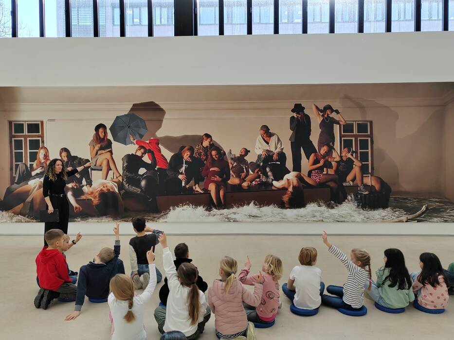 Das Bild zeigt eine Gruppe von Schulkindern, die vor einem großen Kunstwerk sitzen und der Kunstvermittlerin aufmerksam zuhören. Das Kunstwerk selbst ist eine große, detailreiche Fotoarbeit. Die dargestellten Personen sitzen, liegen oder stehen und scheinen in verschiedenen Aktivitäten beschäftigt zu sein. "Untergang der Méduse", 2023/24 von Hannah J. Kohler.