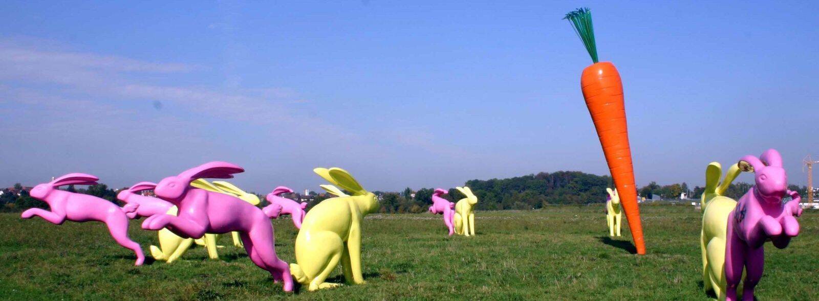 Auf einer grünen Wiese steht eine schräggestellte Karotte. Gelbe Hasen sitzen vor ihr, pinkfarbene Hasen springen fort von ihr.