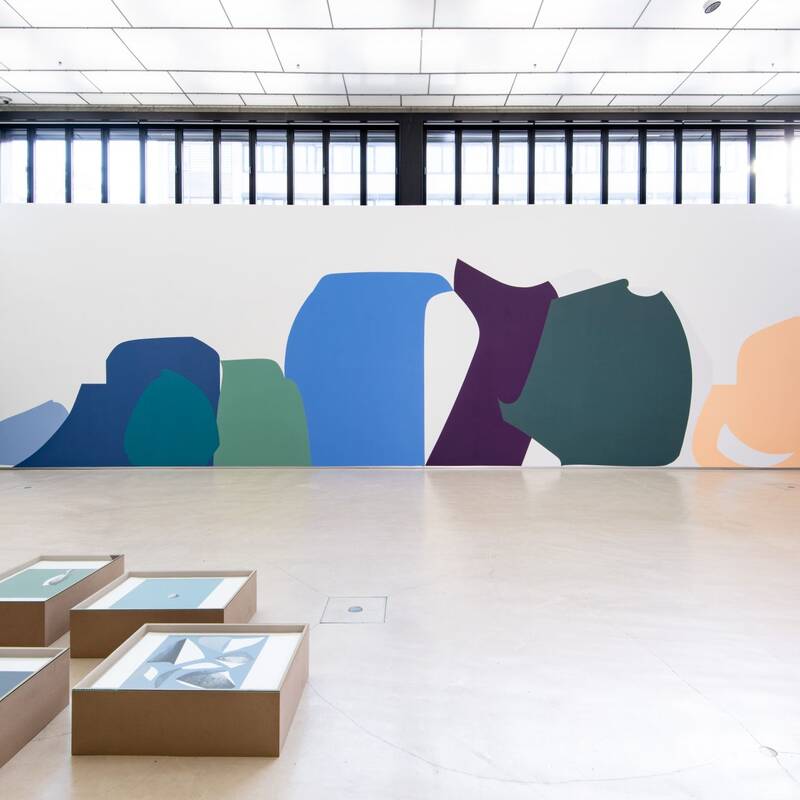 Der Ausstellungsraum zeigt eine große Wandarbeit von farbenfrohen, abstrakten Formen in diversen Blautönen, Grün, Lila und Orange. Im Vordergrund befinden sich mehrere, niedrige Holzschaukästen auf dem Boden.