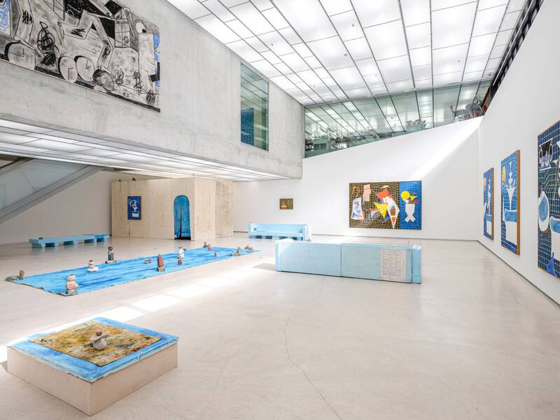 Ausstellungsansicht "Keine Zeit zum Baden" von Jan Zöller. Große Gemälde in Blau- und Gelbtönen dominieren die Wände. Auf dem Boden ist eine Installation zu sehen: Eine blaue Plane, auf dem kleine Steinskulpturen angeordnet sind. Im Vordergrund steht ein quadratisches Objekt mit blau, gelber Oberfläche, auf der wiederum vier Steine aufeinander gestapelt sind.