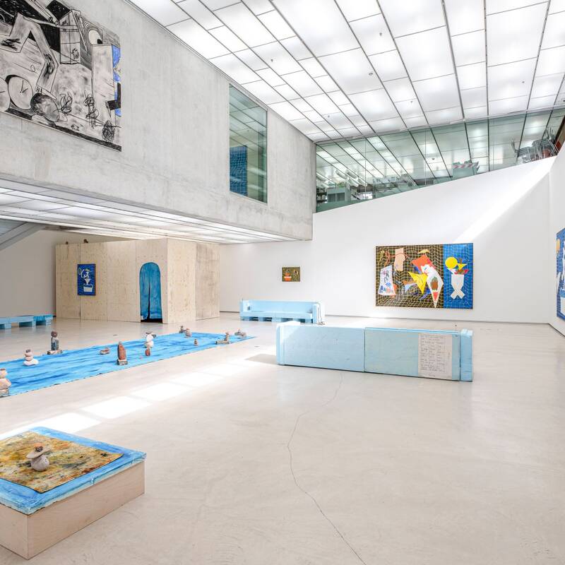 Ausstellungsansicht "Keine Zeit zum Baden" von Jan Zöller. Große Gemälde in Blau- und Gelbtönen dominieren die Wände. Auf dem Boden ist eine Installation zu sehen: Eine blaue Plane, auf dem kleine Steinskulpturen angeordnet sind. Im Vordergrund steht ein quadratisches Objekt mit blau, gelber Oberfläche, auf der wiederum vier Steine aufeinander gestapelt sind.