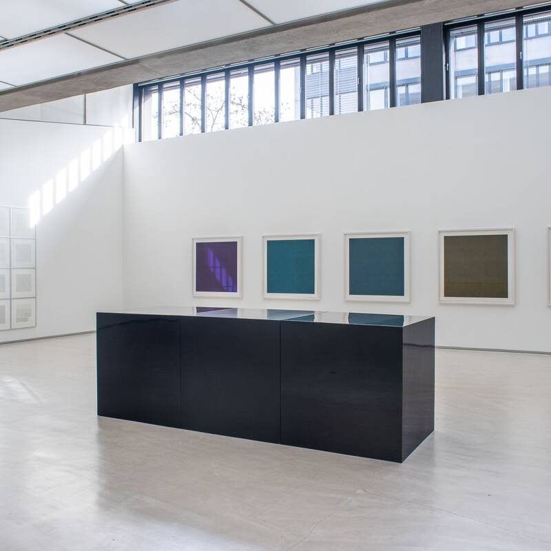 Ausstellungsansicht "Steinreich" von Sol LeWitt. An den Wänden hängen mehrere quadratische Bilder mit einfarbigen Flächen in verschiedenen Farbtönen (Violett, Blau, Grün, Rot). Ein größeres, rasterartig gegliedertes Bild mit hellgrauen Farbnuancen befindet sich an einer Seitenwand. Im Raum steht ein niedriger, schwarzer Kubus.
