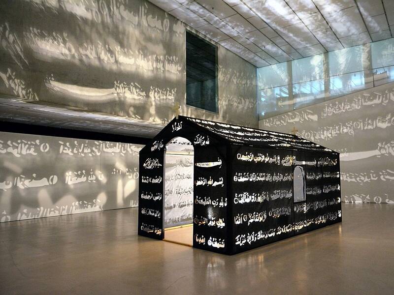 Ausstellungsansicht der Installation "Islamic Chapel" von Nezaket Ekici. In der Mitte des Raumes steht eine Kapellenähnliche Installation. In dessen Wände aus schwarzem Stoff sind arabische Schriftzeichen gestanzt, die durch die Beleuchtung im Inneren der Installation in den Galerieraum projiziert werden.
