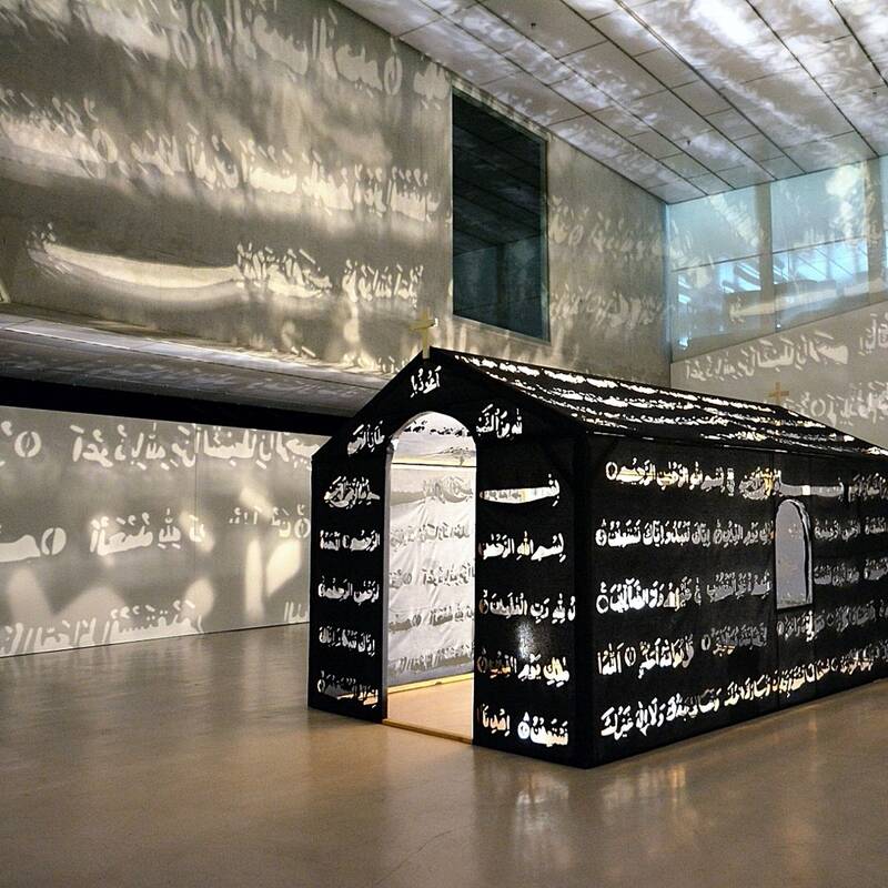 Ausstellungsansicht der Installation "Islamic Chapel" von Nezaket Ekici. In der Mitte des Raumes steht eine Kapellenähnliche Installation. In dessen Wände aus schwarzem Stoff sind arabische Schriftzeichen gestanzt, die durch die Beleuchtung im Inneren der Installation in den Galerieraum projiziert werden.