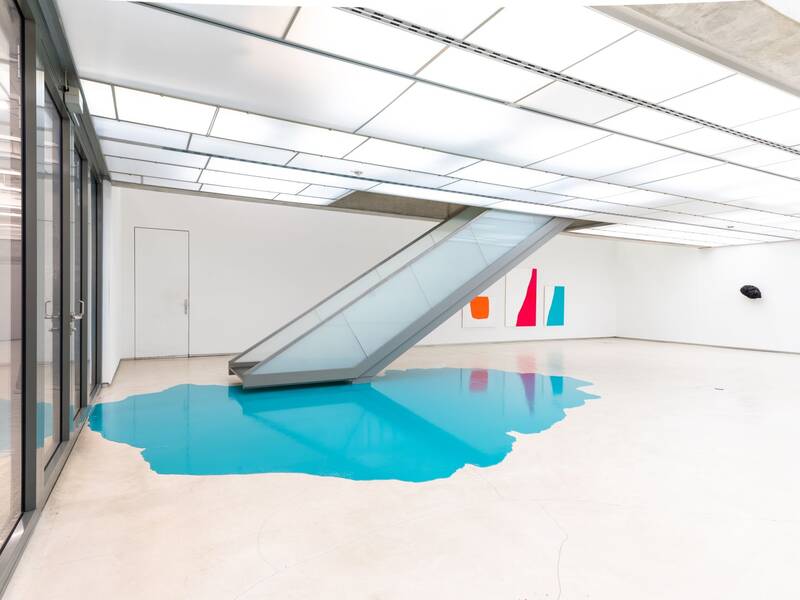 Ausstellungsansicht "Nicht eintönig" von Holder, Lutzeyer und Splitt. Das Bild zeigt eine Treppe, die in einen türkisfarbenen Farbfleck auf dem Boden mündet. An den Wänden hängen abstrakte Gemälde in kräftigen Farben.