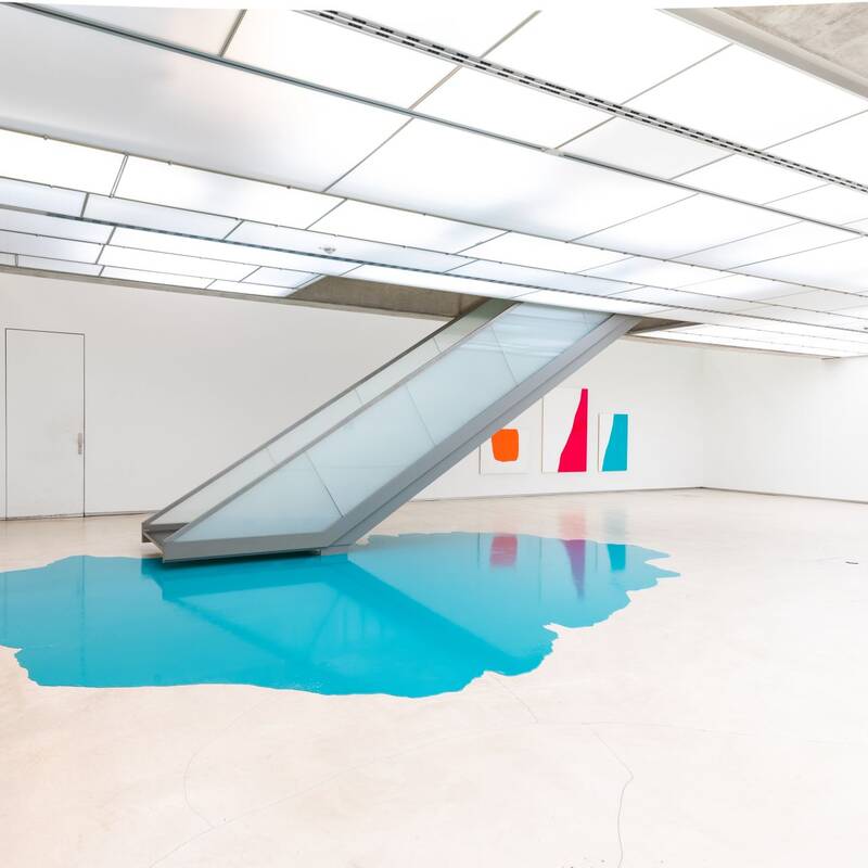 Ausstellungsansicht "Nicht eintönig" von Holder, Lutzeyer und Splitt. Das Bild zeigt eine Treppe, die in einen türkisfarbenen Farbfleck auf dem Boden mündet. An den Wänden hängen abstrakte Gemälde in kräftigen Farben.