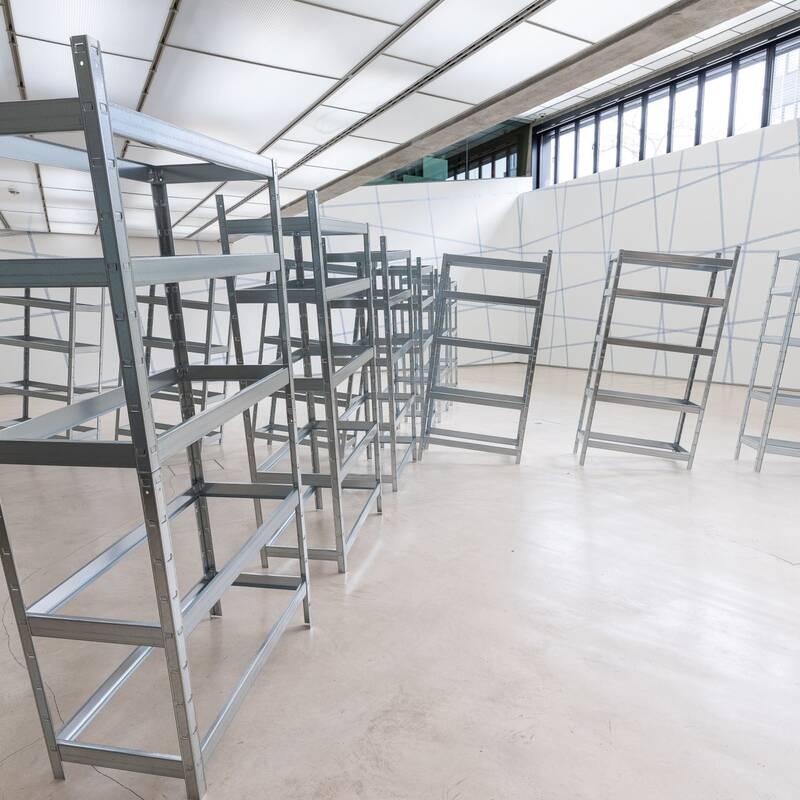 Ausstellungsansicht "Kippmomente" von ststs. Im Raum stehen viele Schwerlastregale, die zu kippen drohen. Die Wände sind mit blauem Klebeband gerastert.