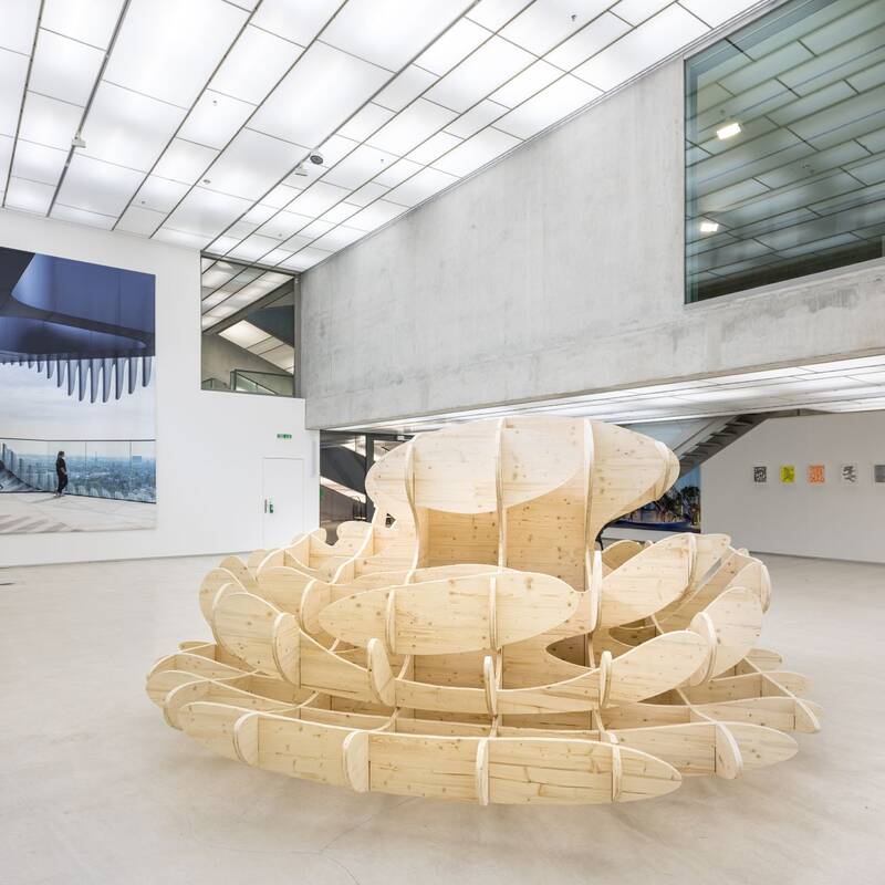 Ausstellungsansicht "Maßstäbe" von J. Mayer H. Eine große, organisch geformte Holzskulptur steht prominent im Raum. Links hängen drei Bildschirme an der Wand. Ein großes, wandflächiges Plakat zeigt eine moderne, städtische Architektur.