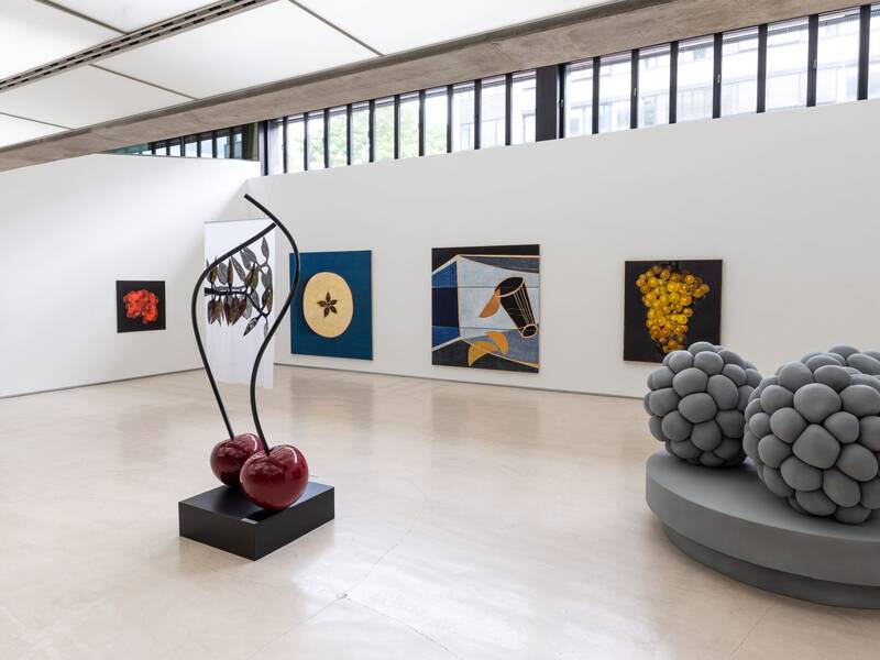 Ausstellungsansicht "Tutti Frutti" der Kunststiftung Rainer Wild. Die Wände zieren große und kleine Bildformate zum Thema Früchte. Zwei große, glänzende Kirschen mit schwarzen Stielen stehen auf einem schwarzen, quadratischen Sockel. Zwei überdimensionale, graue Brombeeren liegen auf einem ovalen, grauen Sockel.