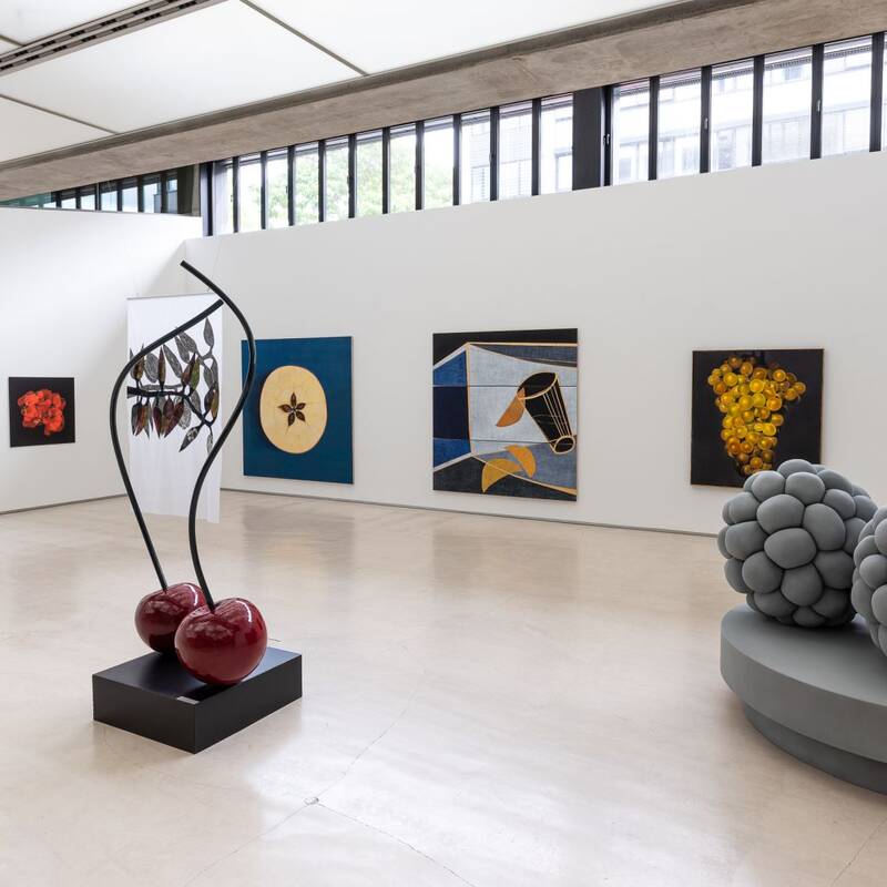 Ausstellungsansicht "Tutti Frutti" der Kunststiftung Rainer Wild. Die Wände zieren große und kleine Bildformate zum Thema Früchte. Zwei große, glänzende Kirschen mit schwarzen Stielen stehen auf einem schwarzen, quadratischen Sockel. Zwei überdimensionale, graue Brombeeren liegen auf einem ovalen, grauen Sockel.