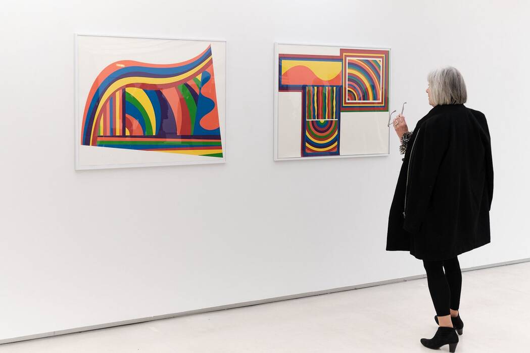 Ausstellung Sol LeWitt: Eine Besucherin betrachtet zwei gerahmte Bilder, die Linien, Bögen und Wellen mit kräftigen bunten Farben zeigen.